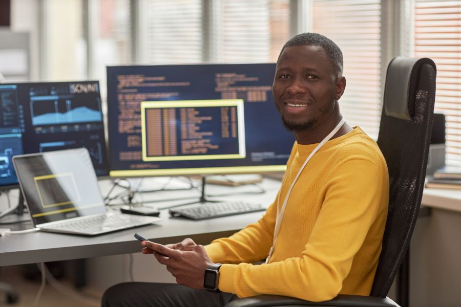 Portrait of smiling black man looking at camera while sitting at workplace in software development company against computer screens with code